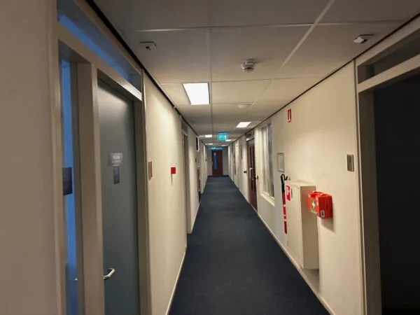 Lang verlichte gang in een kantoorpand aan de Van Bleyswijckstraat met deuren, een EHBO-doos en een brandblusser aan de muur.